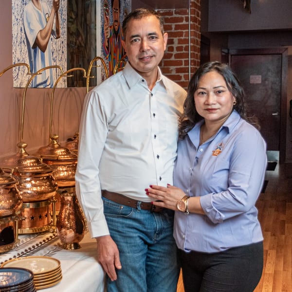 Bir Kitchen owners, a man and woman, stand proudly beside a table with Indian serving dishes.