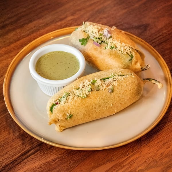 Two golden-fried chili pakoras, garnished with herbs and sev, served with green chutney on a plate.