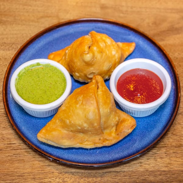 Two golden-brown samosas on a blue plate, served with green chutney and red sauce.