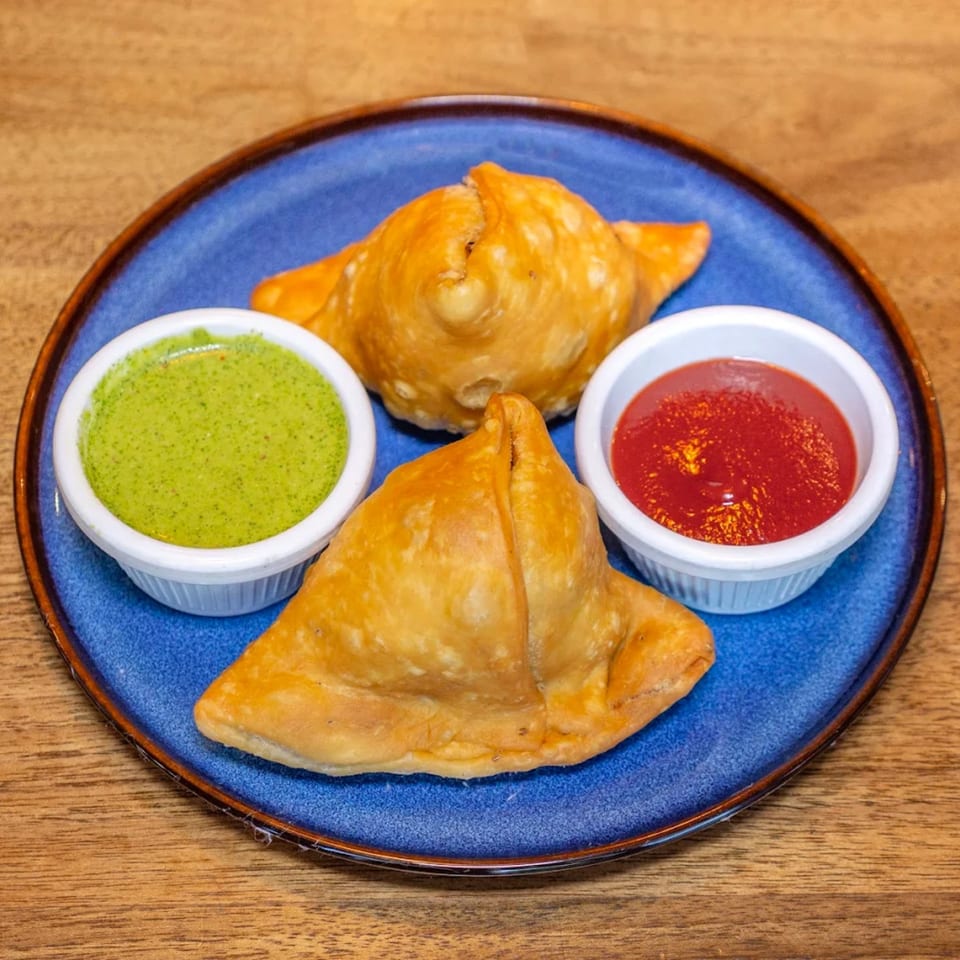 Two golden-brown samosas on a blue plate, served with green chutney and red sauce.