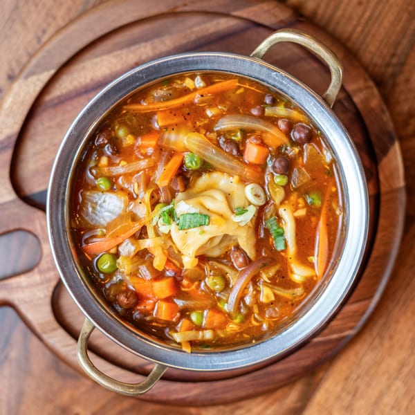A metal bowl of Indian soup with vegetables, chickpeas, and dumplings on a wooden board.