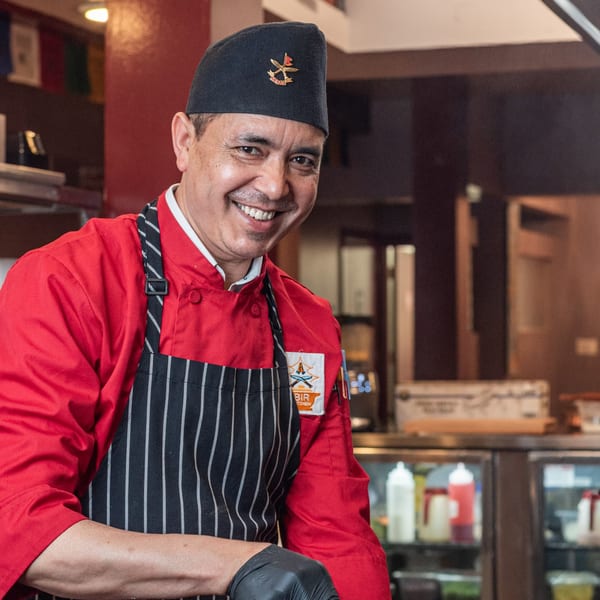 Smiling Bir Kitchen chef in a red uniform and black apron, wearing a black hat with a Nepal pin.