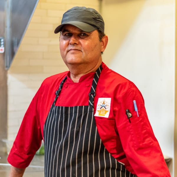 Bir Kitchen chef in a red uniform and striped apron, wearing a dark cap, looking at the camera.
