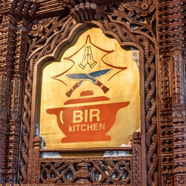 Bir Kitchen's logo, featuring a red cooking pot, crossed knives, and praying hands, framed by ornate wood carvings.