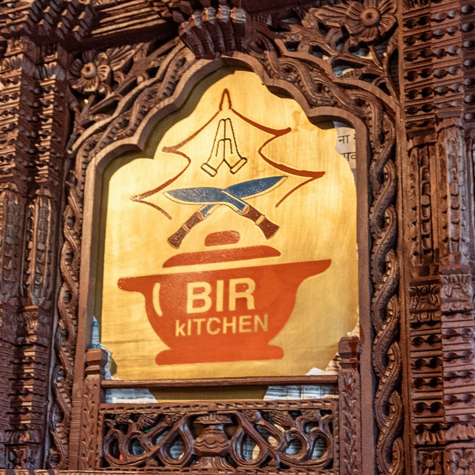 Bir Kitchen's logo, featuring a red cooking pot, crossed knives, and praying hands, framed by ornate wood carvings.