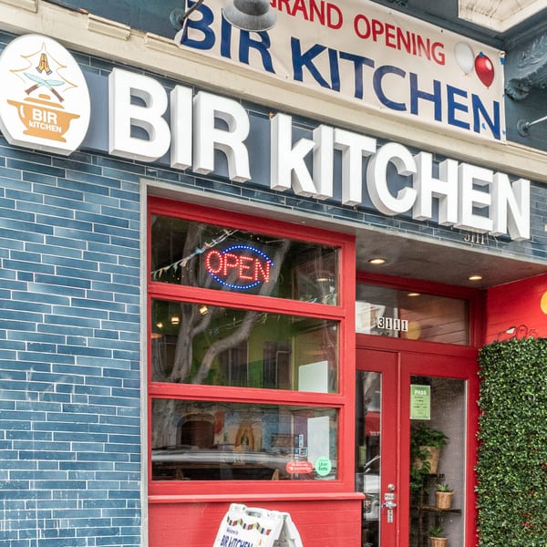 Bir Kitchen's storefront with a blue brick facade, red trim, an "OPEN" sign, and a grand opening banner.