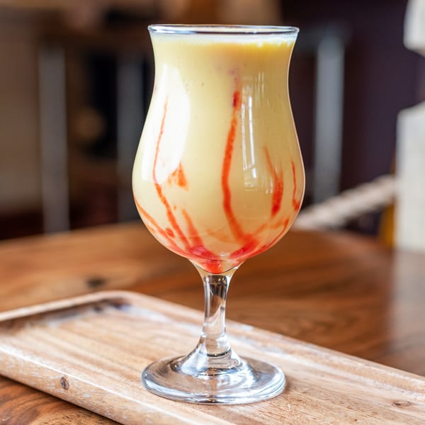 A tall glass of mango lassi with red syrup swirls, on a wooden tray at Bir Kitchen.
